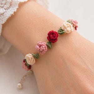 Könnte beinhalten: Ein Häkelarmband mit floralem Design. Das Armband hat rote, rosa und cremefarbene Rosen mit grünen Blättern. Es befindet sich auf einer beigefarbenen Kordel und hat eine Perlenperle am Ende.