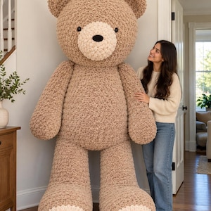 Peut inclure: Un ours en peluche géant crocheté de couleur brun clair avec un museau et des coussinets de couleur crème. L'ours a des yeux ronds foncés et un nez noir. Le texte en haut indique : FINISHED SIZE: APPROX. 8 FT TALL. Il présente une texture en laine.