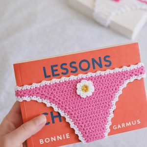 May include: A pink crochet book cover shaped like underwear, with white trim and a small flower embellishment. The book has an orange cover with the title "LESSONS" in blue. A matching crochet item is in the background.