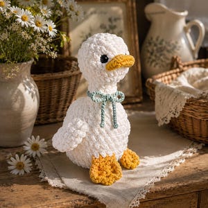 May include: A white crocheted duck plush toy with a textured, fuzzy yarn finish. The duck features a bright yellow beak and matching yellow webbed feet. A small, light blue crocheted bow is tied around its neck, and it has dark, round eyes.
