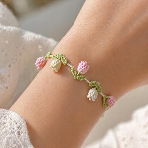 Può includere: Un delicato braccialetto all'uncinetto con dettagli floreali rosa e bianchi e foglie verdi. Il braccialetto è indossato su un polso, mettendo in risalto il design floreale. Un accessorio affascinante.