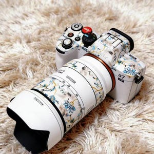 Può includere: Una fotocamera bianca con un motivo floreale sul corpo e sull'obiettivo. L'obiettivo ha un anello bianco e marrone. La fotocamera è appoggiata su una superficie soffice color crema. L'obiettivo ha il marchio "TAMRON".