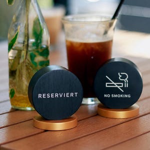 Könnte beinhalten: Zwei runde schwarze Holzschilder mit goldfarbenen Sockeln. Ein Schild trägt den weißen Schriftzug "RESERVIERT" und das andere zeigt ein weißes "NO SMOKING"-Symbol mit dem entsprechenden Text darunter, platziert auf einer Holzlattenoberfläche.