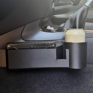 May include: A black plastic car interior organiser featuring a rectangular storage compartment and a cylindrical cup holder. The organiser is mounted to the side of the centre console, holding a metallic beverage can and a black rectangular object.