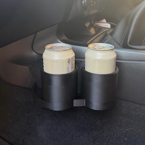 May include: A black dual cup holder insert designed for a vehicle console, holding two light-coloured aluminium beverage cans. The holder features a cylindrical shape with a smooth finish and a connecting bridge between the two circular compartments.