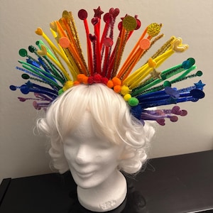Könnte beinhalten: Ein skurriles Kopfstück in Regenbogenfarben, das aus Pfeifenreinigern und dekorativen Formen besteht. Das Kopfstück ist mit roten, orangen, gelben, grünen, blauen und violetten Elementen verziert, darunter Herzen, Sterne und Kreise. Es sitzt auf einem weißen Schaufensterkopf.