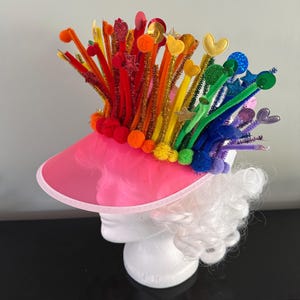 Könnte beinhalten: Ein pinkfarbenes Visier mit einem Regenbogen aus bunten Pfeifenreinigern, Pompons und Stern- und Herzdekorationen. Das Visier sitzt auf einem weißen Perückenkopf und schafft ein skurriles und festliches Accessoire.