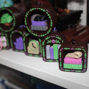 Puede incluir: Una colección de adornos decorativos marrones en miniatura, cada uno con un diseño interior único. Algunos presentan un gato, libros, un reloj o una calavera. Los adornos están decorados con detalles verdes y morados y están dispuestos sobre una superficie blanca.