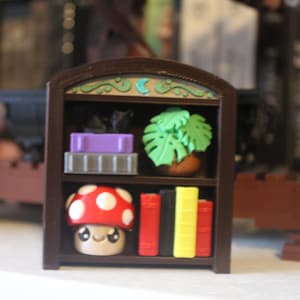 Può includere: Una piccola libreria marrone è piena di oggetti decorativi. Presenta una figura di fungo rosso e bianco, piccoli libri, una pianta in vaso e altri oggetti colorati. La parte superiore della libreria ha un design decorativo.