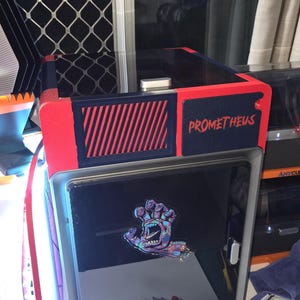 Könnte beinhalten: Ein 3D-Drucker in Schwarz und Rot. Auf der Oberseite steht das Wort "PROMETHEUS" in Rot. Auf der Vorderseite befindet sich eine Grafik einer Monsterhand. Die Worte "ANYCUBIC KOBRA S1" sind auf der Vorderseite des Druckers zu sehen.