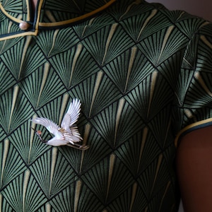 May include: A green dress with a geometric fan pattern and a white crane brooch. The dress has a mandarin collar and yellow trim. The brooch is a detailed sculpture of a crane in flight, adding a touch of elegance to the outfit.