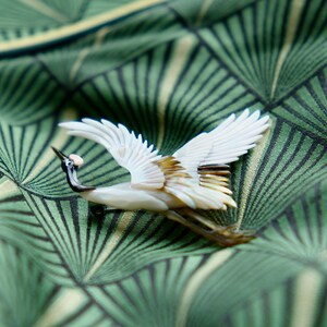 May include: A detailed crane brooch with outstretched wings, crafted from white and iridescent materials. The bird has a black beak and neck, with a small pink accent. The brooch is set against a green patterned fabric.