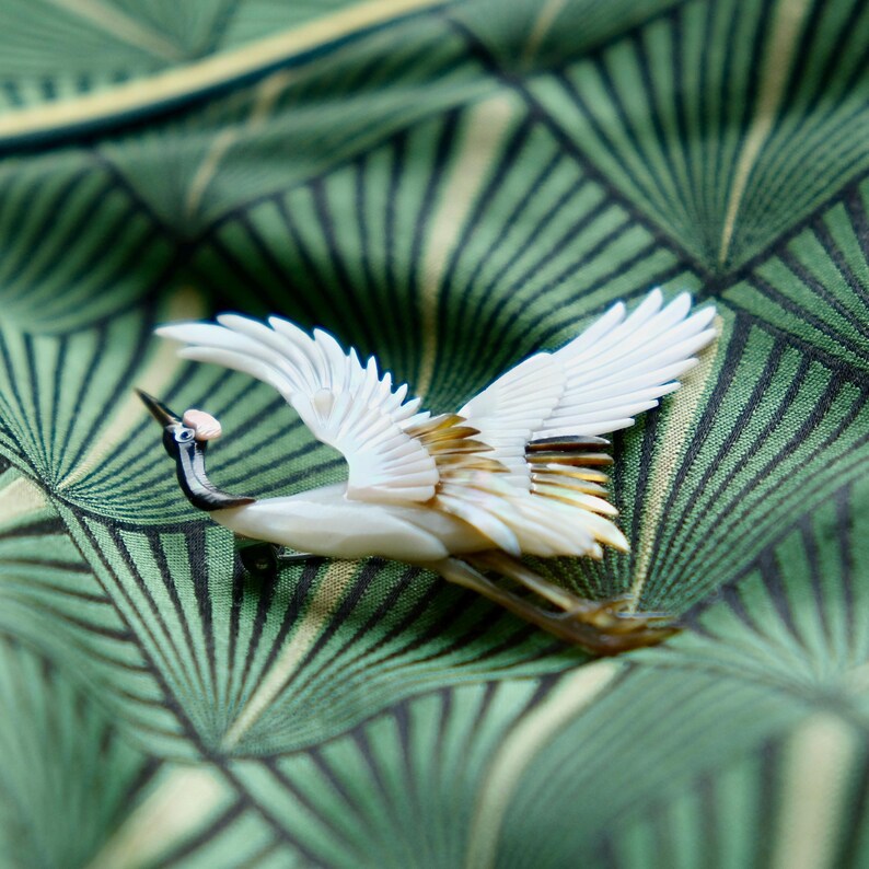 May include: A detailed crane brooch with outstretched wings, crafted from white and iridescent materials. The bird has a black beak and neck, with a small pink accent. The brooch is set against a green patterned fabric.