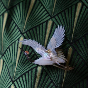 May include: A white and iridescent crane brooch with outstretched wings, featuring a red spot on its head and a dark beak. The bird is set against a green and gold geometric patterned background.