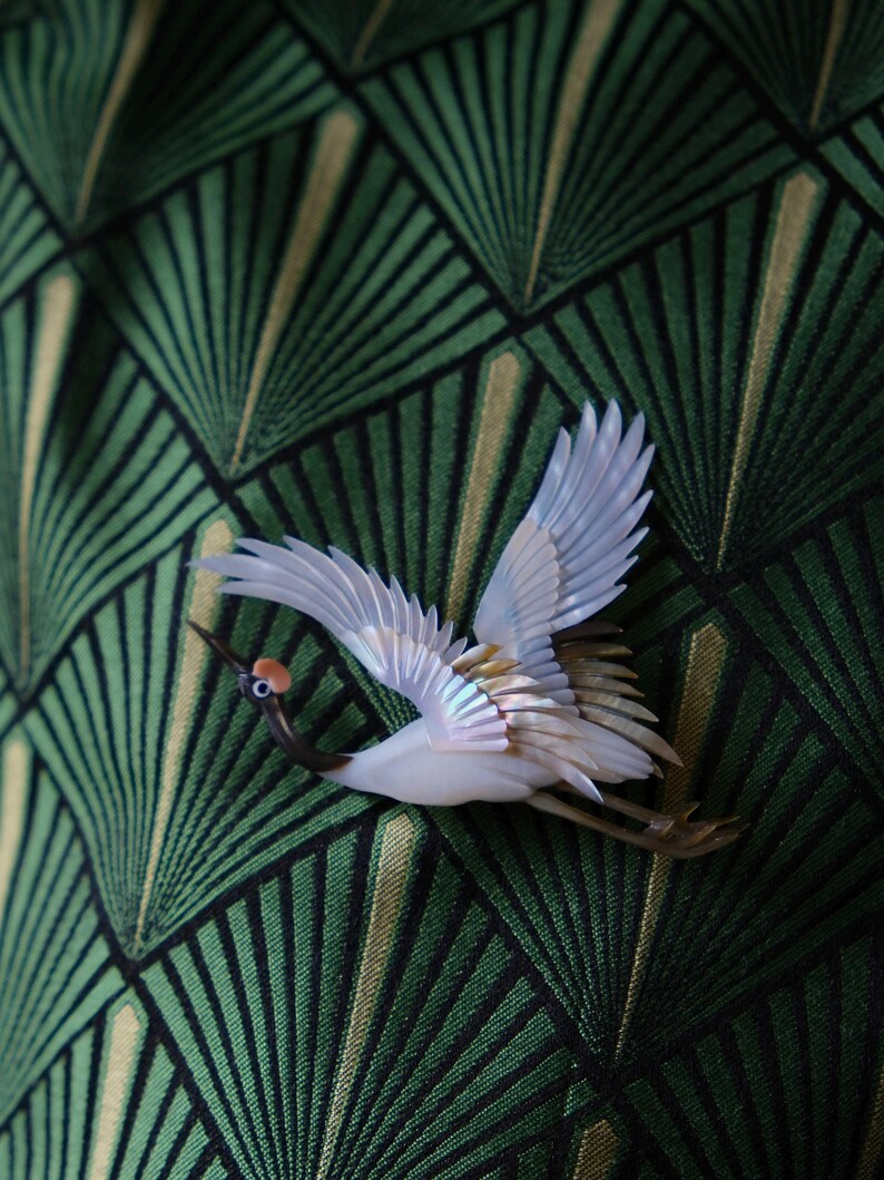May include: A white and iridescent crane brooch with outstretched wings, featuring a red spot on its head and a dark beak. The bird is set against a green and gold geometric patterned background.
