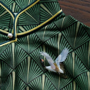 May include: Close-up of a green patterned fabric with a fan-like design. The fabric is trimmed with a yellow and dark green border. A white and brown bird brooch is attached to the fabric. A pearl button is visible.