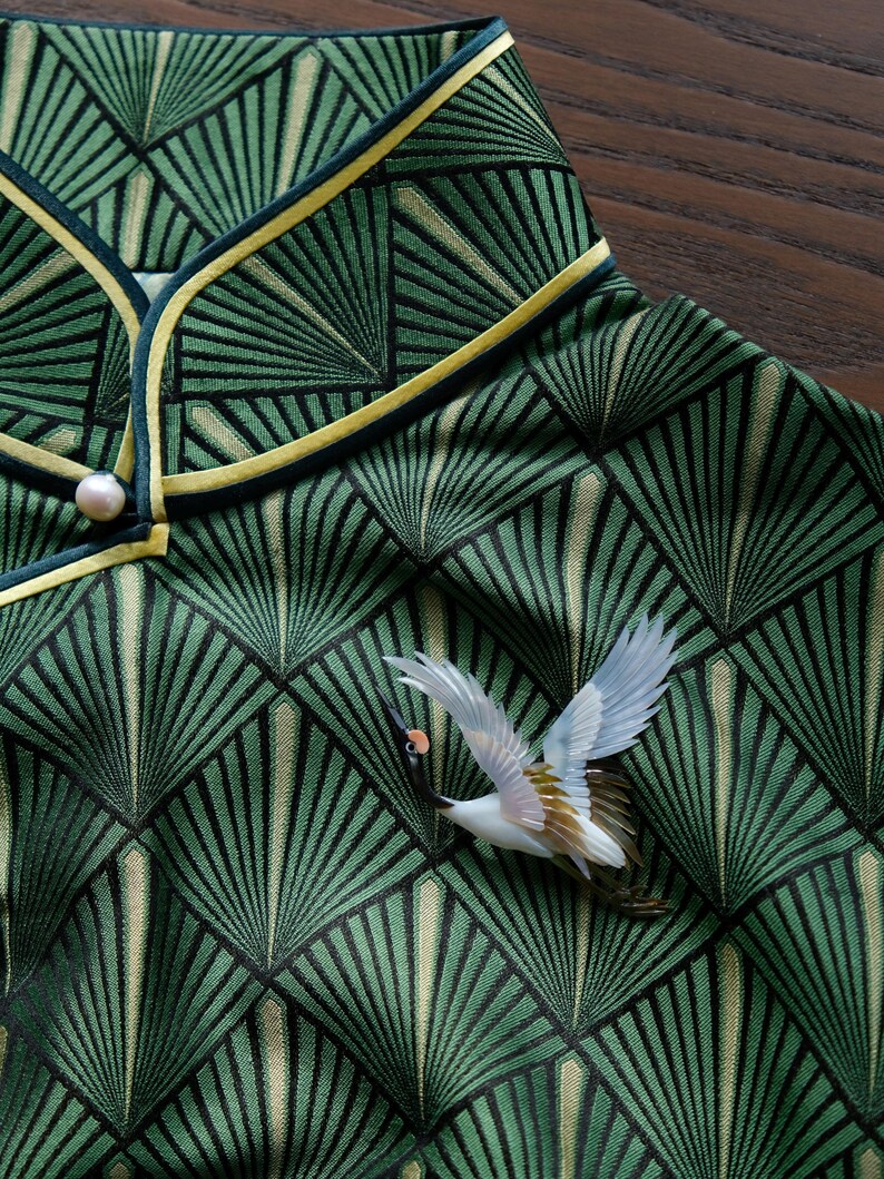 May include: Close-up of a green patterned fabric with a fan-like design. The fabric is trimmed with a yellow and dark green border. A white and brown bird brooch is attached to the fabric. A pearl button is visible.