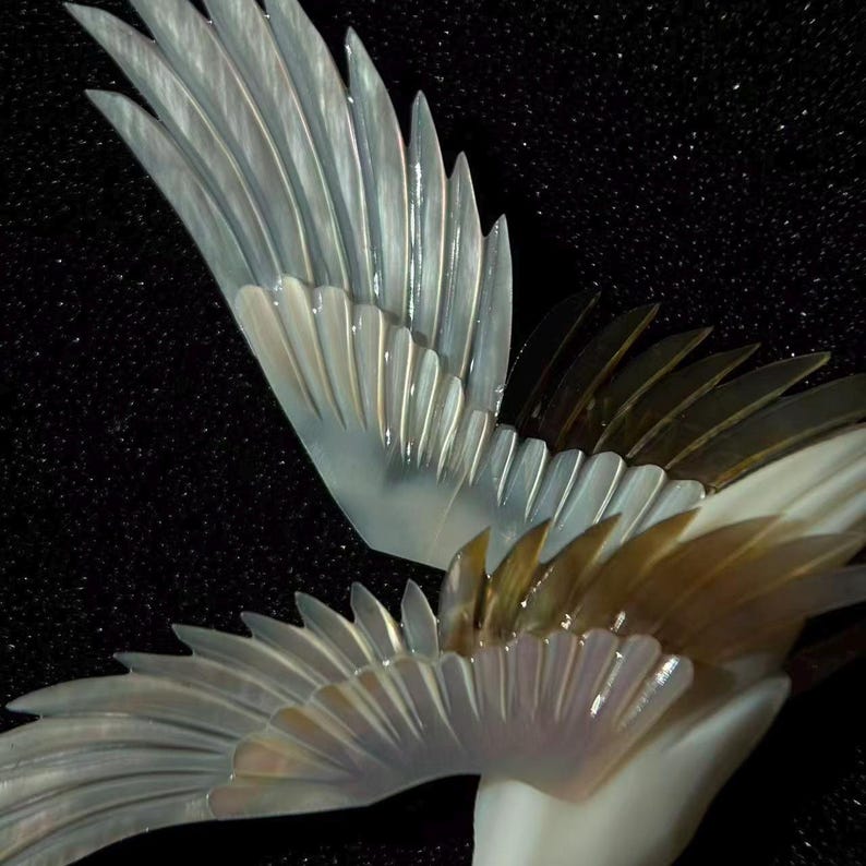 May include: Close-up of a pair of iridescent, stylized wings, possibly a sculpture or decorative element. The wings feature a layered design with a pearlescent sheen, transitioning from white to a golden hue. The background is a dark, textured surface.