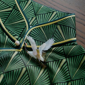 May include: Close-up of a green garment with a geometric fan pattern, accented with yellow trim and a decorative crane brooch. The fabric has a textured appearance, and the brooch adds a touch of elegance.