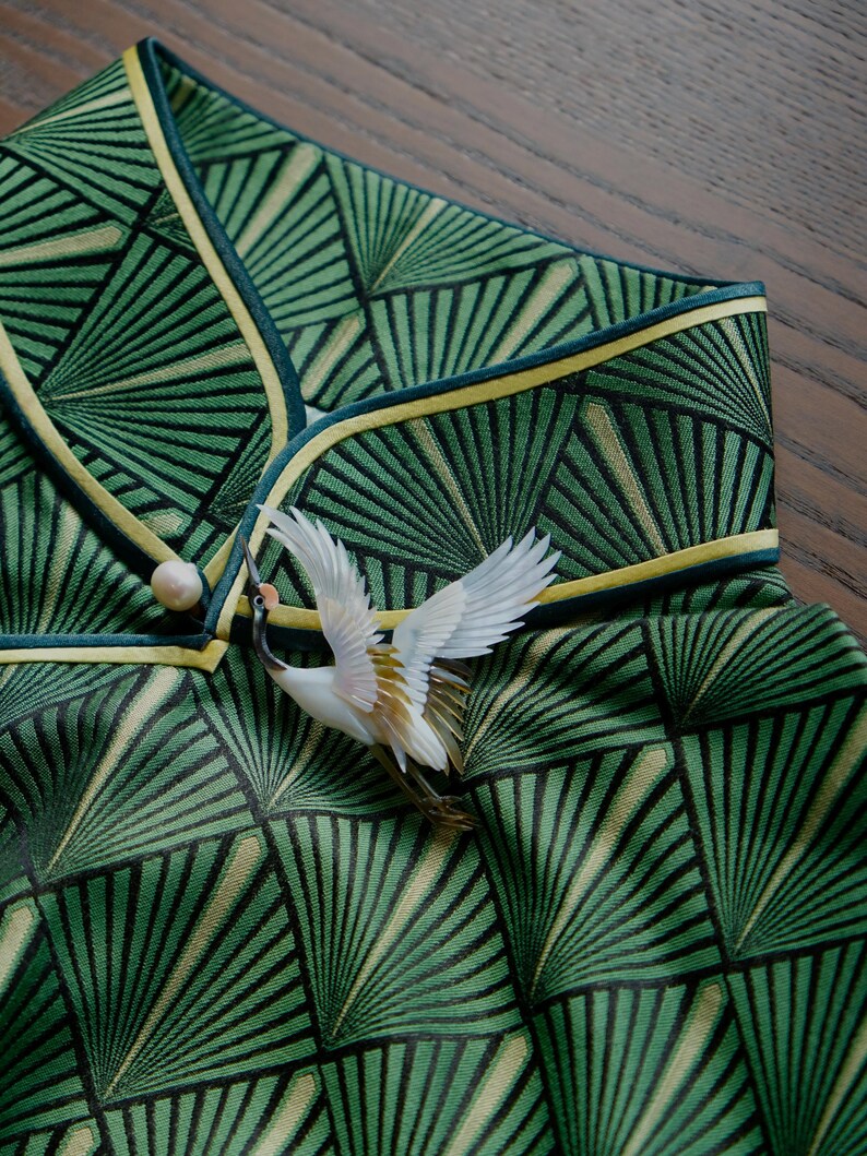 May include: Close-up of a green garment with a geometric fan pattern, accented with yellow trim and a decorative crane brooch. The fabric has a textured appearance, and the brooch adds a touch of elegance.