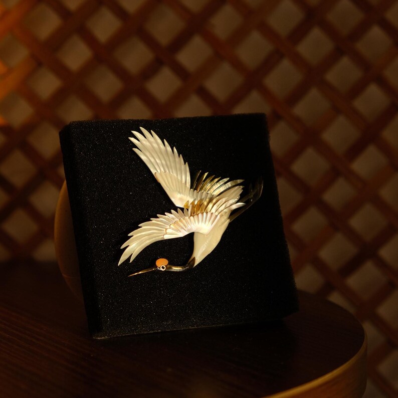 May include: A detailed, elegant crane brooch with outstretched wings. The bird is primarily white with gold accents on the wings and a small orange detail. The brooch is displayed on a black surface, set against a warm, brown background.