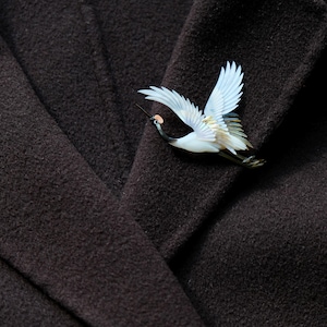 May include: A detailed crane brooch with outstretched wings, featuring white, blue, and tan hues. The bird's beak and legs are black, and it has a small pink accent. The brooch is set against a dark brown fabric, creating a striking contrast.