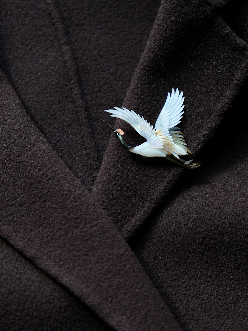 May include: A detailed crane brooch with outstretched wings, featuring white, blue, and tan hues. The bird's beak and legs are black, and it has a small pink accent. The brooch is set against a dark brown fabric, creating a striking contrast.