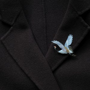 May include: A black coat with a decorative bird pin. The pin features a crane in flight, with blue wings and detailed plumage. The coat's fabric has a subtle texture.