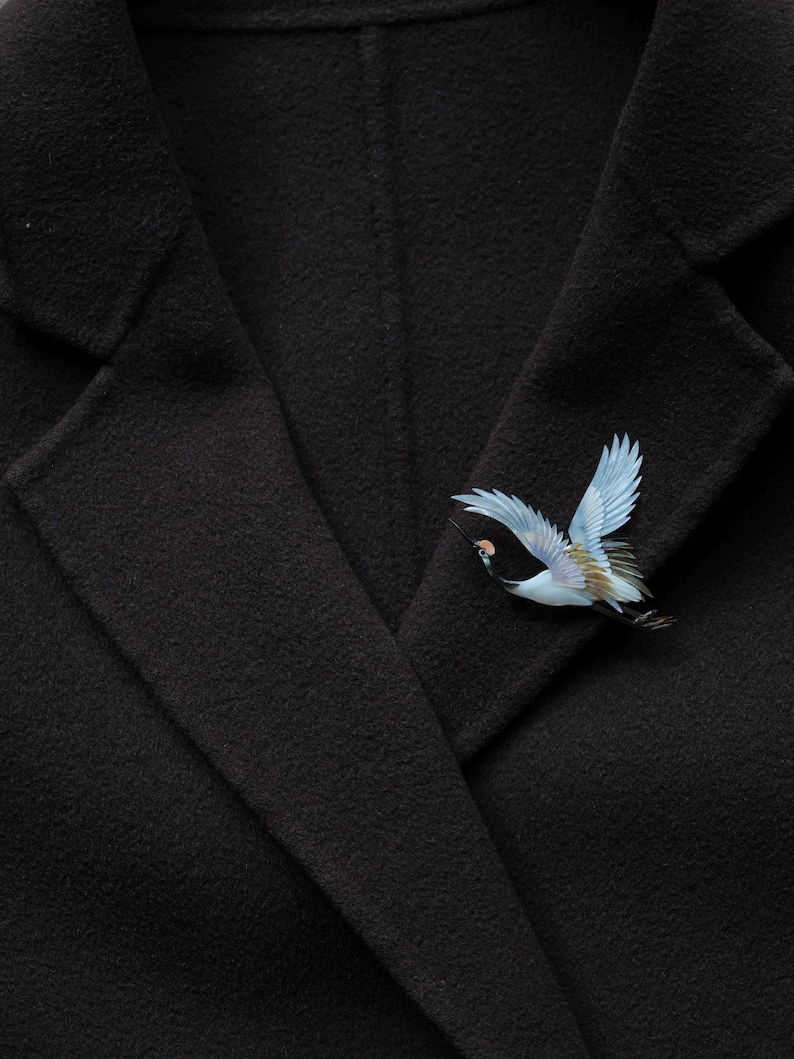May include: A black coat with a decorative bird pin. The pin features a crane in flight, with blue wings and detailed plumage. The coat's fabric has a subtle texture.
