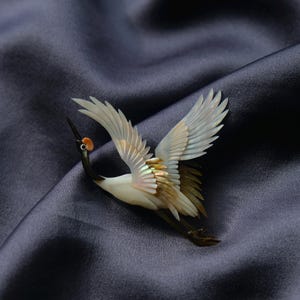 May include: A detailed crane brooch with outstretched wings, crafted from iridescent materials. The bird features a white body, black neck, and a small orange head. The brooch is set against a dark blue, textured background.