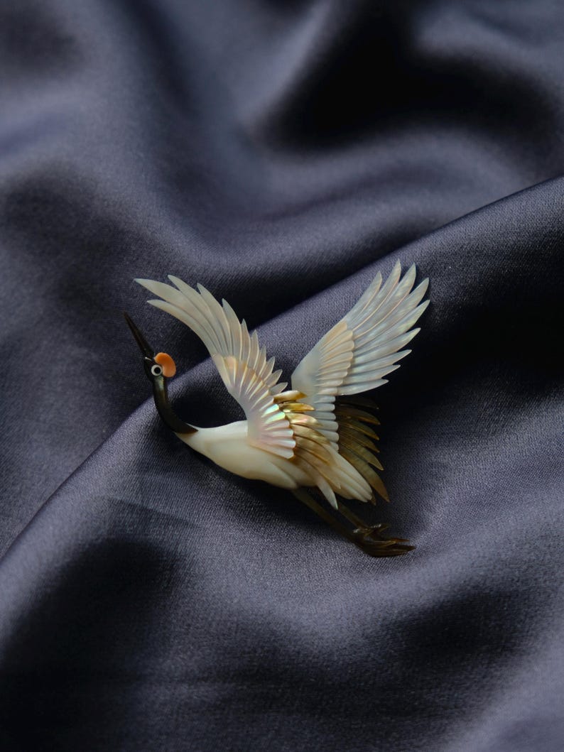 May include: A detailed crane brooch with outstretched wings, crafted from iridescent materials. The bird features a white body, black neck, and a small orange head. The brooch is set against a dark blue, textured background.