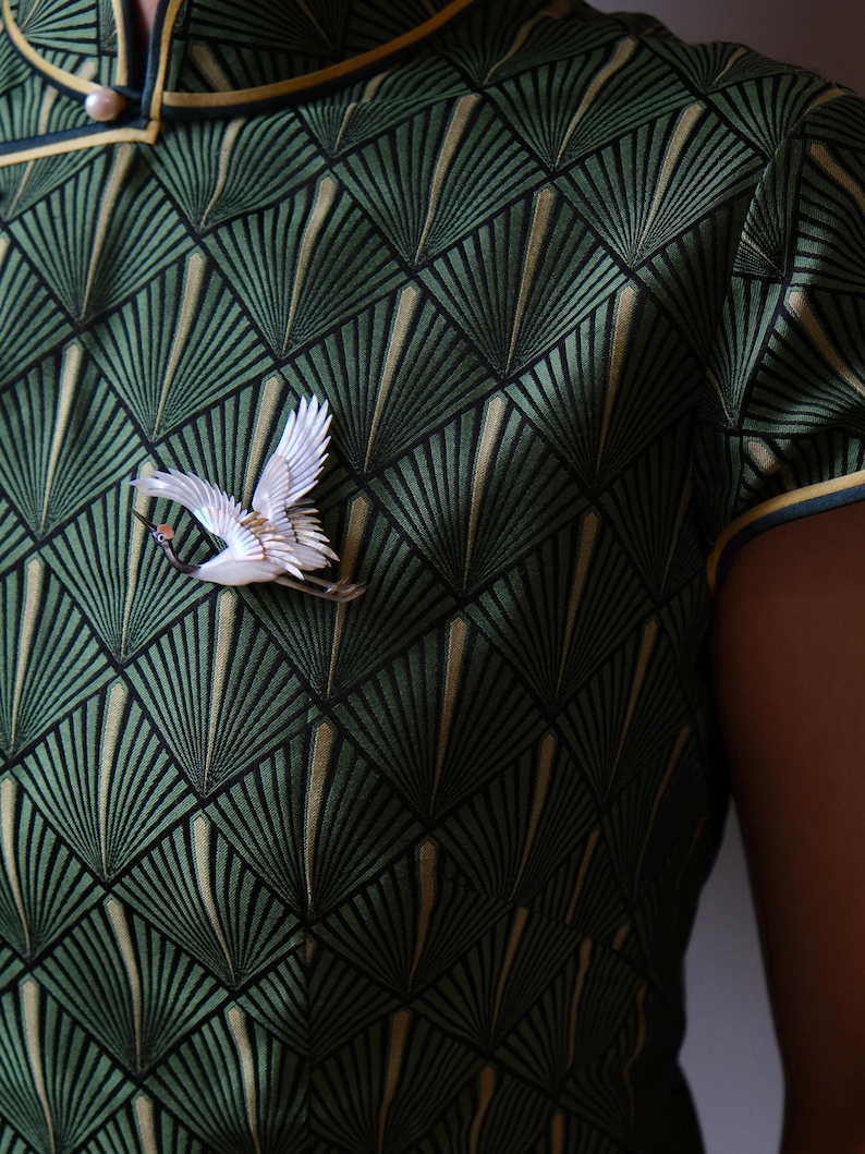 May include: A green dress with a geometric fan pattern and a white crane brooch. The dress has a mandarin collar and yellow trim. The brooch is a detailed sculpture of a crane in flight, adding a touch of elegance to the outfit.