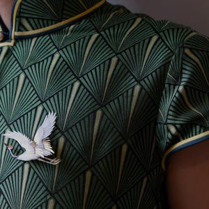 May include: Green patterned top with a white crane brooch. The top features a geometric fan design in shades of green and gold, with a mandarin collar and yellow trim. A white crane brooch is pinned on the front.