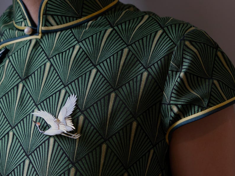May include: Green patterned top with a white crane brooch. The top features a geometric fan design in shades of green and gold, with a mandarin collar and yellow trim. A white crane brooch is pinned on the front.