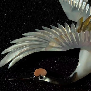 May include: Close-up of a decorative bird with iridescent wings and a long, slender beak. The wings are detailed with a layered, feathery design, and the bird's head features a small, rounded crest. The background is a dark, textured surface.