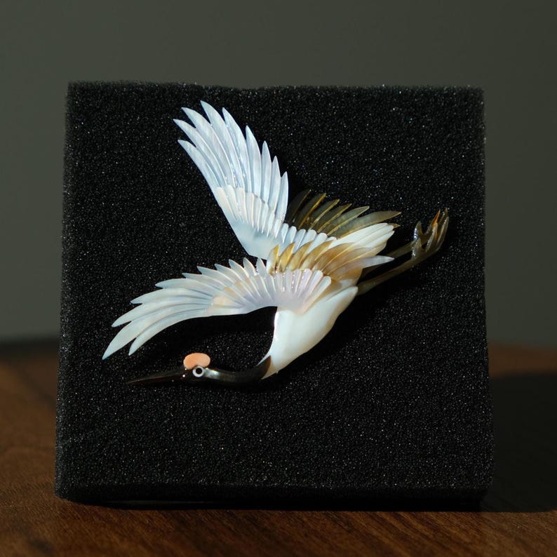 May include: A detailed sculpture of a crane in flight, featuring white, gold, and black colors. The bird's wings are spread, showcasing intricate feather details. The crane has a black beak and a small, orange-colored head crest. The sculpture is displayed on a black, textured surface.
