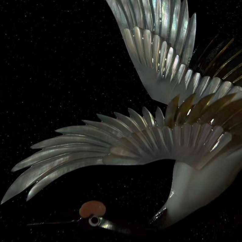 May include: Close-up of a decorative bird with iridescent wings and a black background. The wings are detailed with a layered, textured design, showcasing shades of silver, white, and brown. The bird's body is white, and its beak is black.