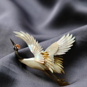 May include: A crane brooch with outstretched wings, crafted from iridescent materials. The bird features a white body, black neck, and touches of gold and pink. The brooch is set against a gray, textured background.