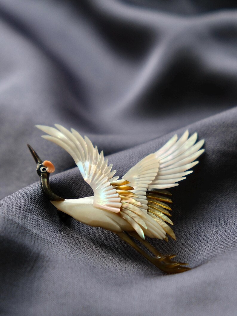 May include: A crane brooch with outstretched wings, crafted from iridescent materials. The bird features a white body, black neck, and touches of gold and pink. The brooch is set against a gray, textured background.