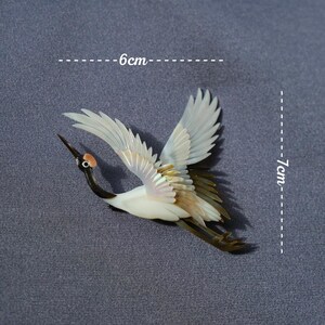 May include: A crane figurine with outstretched wings, measuring approximately 6 cm long and 7 cm wide. The bird features a white body, black neck, and detailed wing feathers with brown and iridescent accents. The crane is depicted in flight.