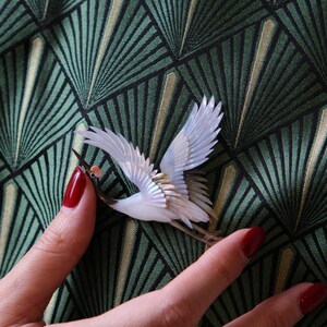 May include: A detailed crane brooch with white wings and a brown beak. The bird is set against a green and black geometric patterned background. The brooch is held by a person with red nail polish.
