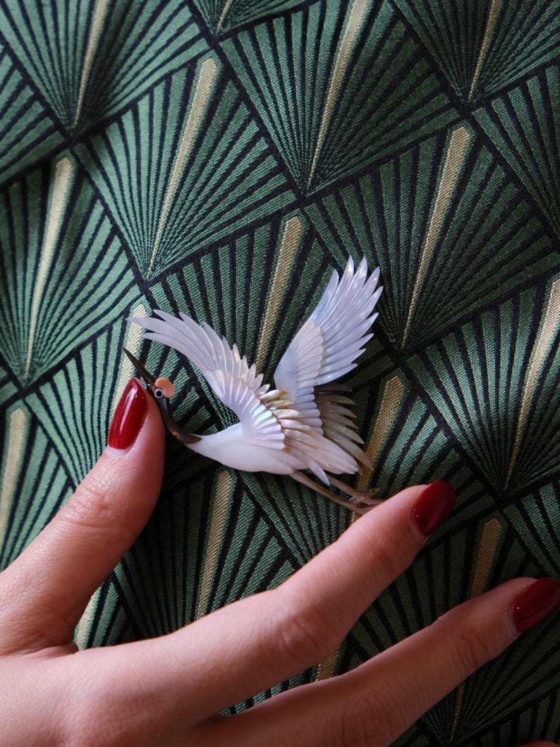 May include: A detailed crane brooch with white wings and a brown beak. The bird is set against a green and black geometric patterned background. The brooch is held by a person with red nail polish.