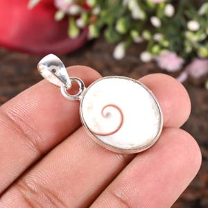 Peut inclure: Pendentif ovale avec un motif en spirale blanc et marron serti dans un métal argenté. Le pendentif a une boucle en haut pour une chaîne. L'arrière-plan est une vue floue d'une surface en bois et de verdure.