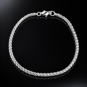 Puede incluir: Una pulsera de cadena tipo trigo en tono plateado con un acabado metálico pulido y un cierre de mosquetón seguro. La cadena está construida con eslabones entrelazados complejos que crean una textura redondeada y uniforme en toda la pieza.