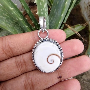Puede incluir: Un colgante de plata con una concha de ojo de Shiva blanca y redonda con un patrón de espiral marrón natural. La concha está engastada en un marco de plata pulida con un borde de cuerda trenzada y una anilla de plata para colgar en cadena.