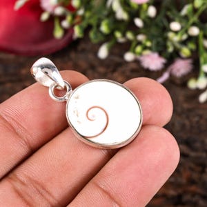 Op de afbeelding: Zilveren hanger met een wit, spiraalvormig ontwerp. De hanger heeft een ronde vorm met een zilveren lus voor een ketting. De spiraal is lichtbruin, tegen een witte achtergrond. Sieraden.