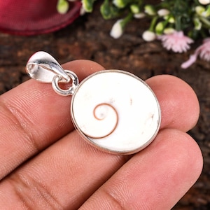 Peut inclure: Pendentif rond avec un fond blanc et un motif en spirale au centre. Le pendentif est encadré d'une monture en métal argenté et possède une bélière argentée pour l'attacher à une chaîne. Le pendentif est tenu dans une main.