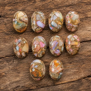 May include: Ten oval-shaped cabochons featuring a mosaic pattern of multicolored stone fragments embedded in a metallic gold matrix. Each polished gemstone displays a unique arrangement of pink, orange, white, and green mineral inclusions.