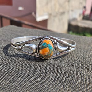 Könnte beinhalten: Ein silbernes Armband mit einer zentralen, ovalen Steinverzierung, die ein marmoriertes Muster in leuchtendem Orange, Türkisblau und Braun aufweist. Das Metallband ist poliert und besitzt geschwungene, offene Verzierungen an beiden Seiten.