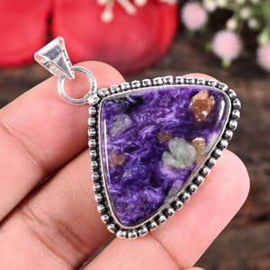 Puede incluir: Un colgante triangular con una fianza plateada. El colgante presenta una piedra púrpura pulida con patrones arremolinados y motas marrones y grises. La piedra está engastada en un marco plateado con cuentas.